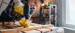 A woman using a power drill to drive a screw into a piece of lumber.